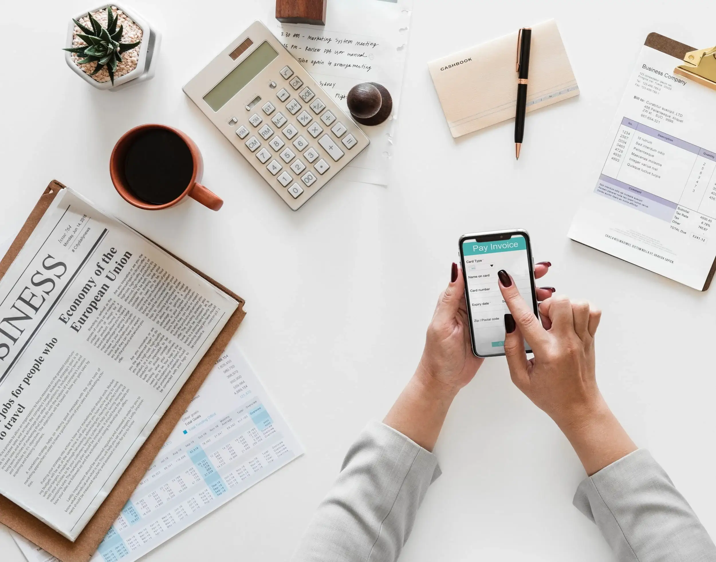 Seen from above, a desk with a calculator, a coffee, a newspaper and hands holding a phone