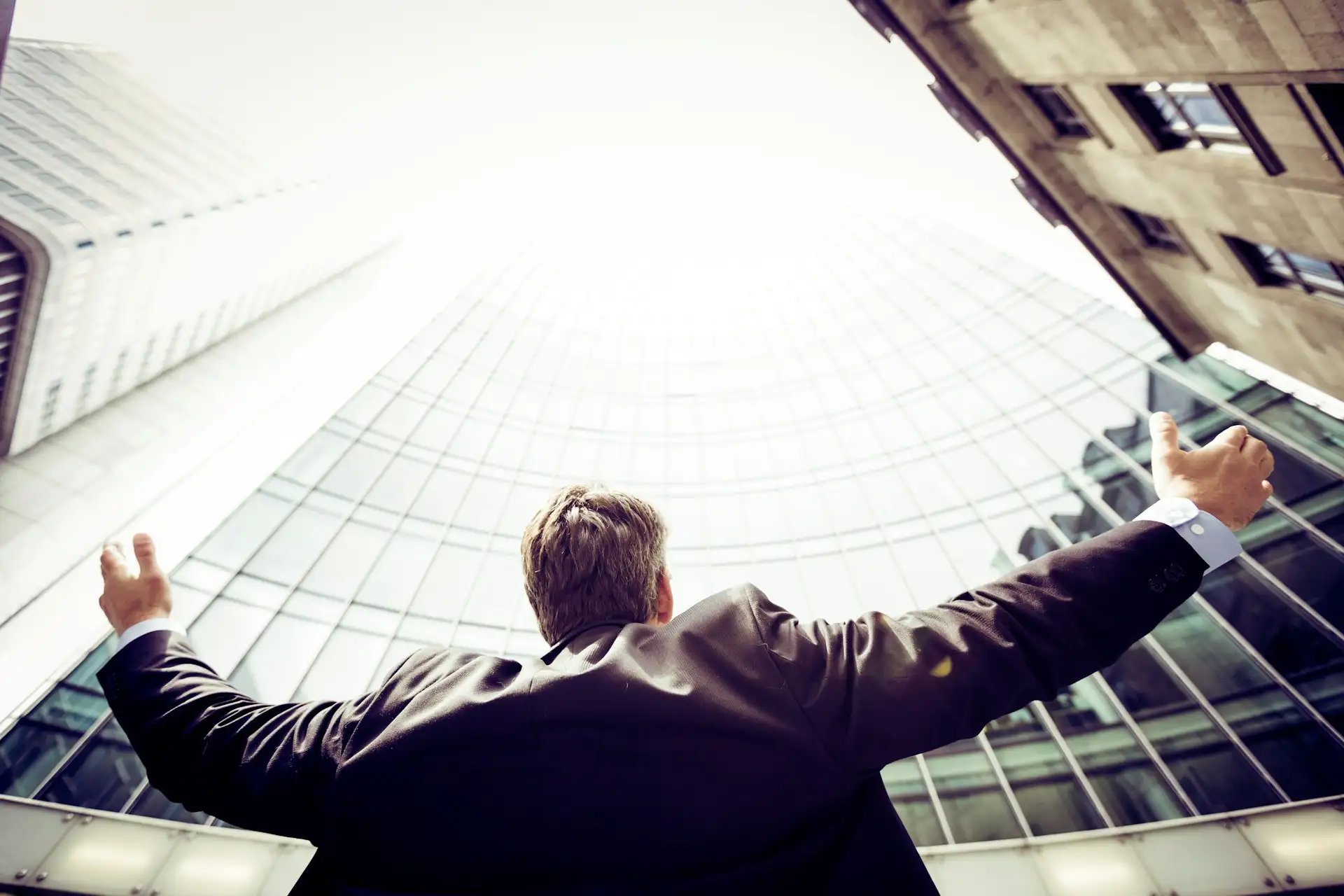 A businessman with his arms raised wide, looking up between glass office towers
